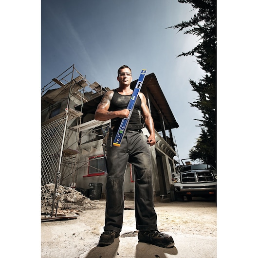 A construction worker in a black tank top and work pants stands outdoors in front of a building under renovation. The worker holds a blue IRWIN level tool. Scaffolding and a pickup truck are visible in the background. The worker's face is blurred for privacy.