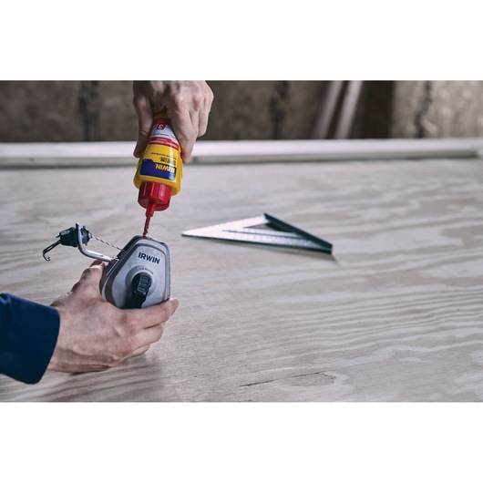Photo of hands filling an IRWIN 1932890 chalk line reel with chalk, metal square on wooden surface in background.