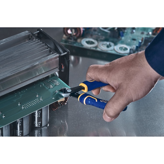 A hand holding an IRWIN blue and yellow wire cutter, cutting wires on an electronic circuit board placed on a metallic surface.