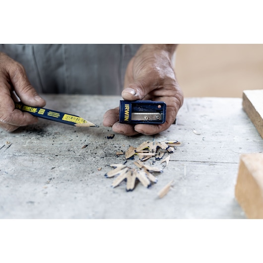 Photo of hands sharpening a blue IRWIN carpenter pencil with IRWIN sharpener 233250, wood shavings on workbench.