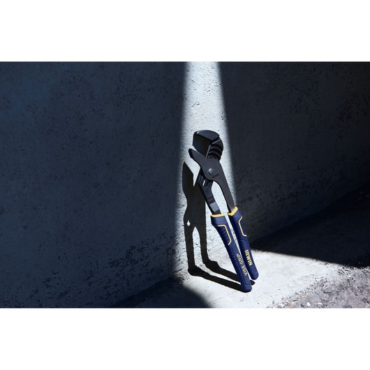An IRWIN branded adjustable plier with blue and yellow handles is standing upright against a textured gray wall, illuminated by a narrow beam of sunlight.