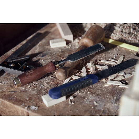 IRWIN 2011491 chisel and rasp with blue handle on a dusty woodworking bench, surrounded by wood shavings.