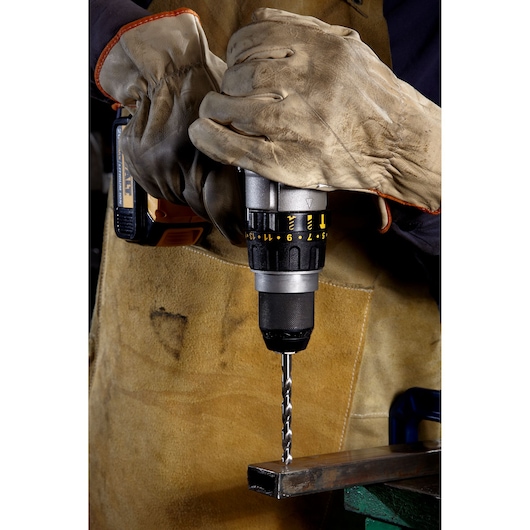 Close-up of gloved hands operating a cordless power drill with a metal drill bit, drilling into a metal surface. The person is wearing a protective brown apron.