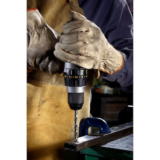 Person wearing protective gloves and an apron is using a power drill to make a hole in a metal bar held in place by a clamp.