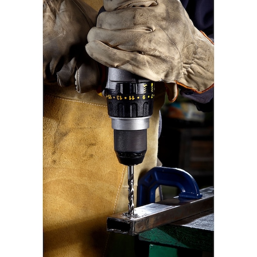 A person wearing protective gloves uses a power drill to make a hole in a metal piece secured with a clamp.