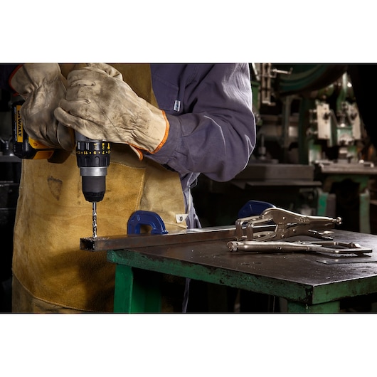 A person wearing gloves and protective clothing is using a cordless drill to work on a metal piece secured with an IRWIN clamp on a workbench. Other metal tools are placed on the table.