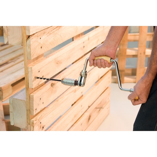 A person using a hand drill with a wooden handle to drill into a wooden pallet.