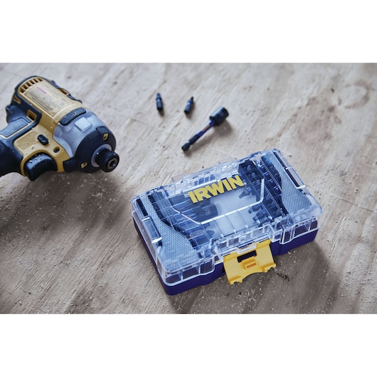 A blue and yellow IRWIN branded plastic case containing tool bits is placed on a wooden surface next to a cordless power drill and several loose bits.