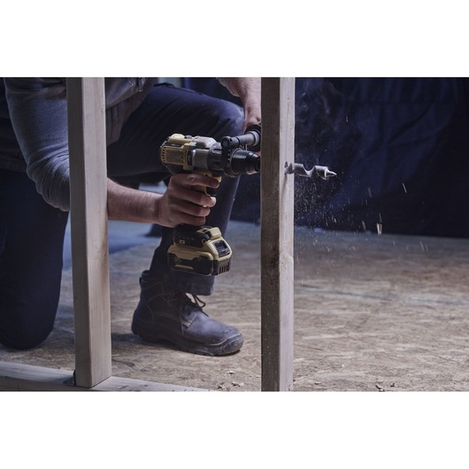 Close-up photo of a person using a DeWalt drill with auger bit to drill through a vertical wooden stud on a construction site.