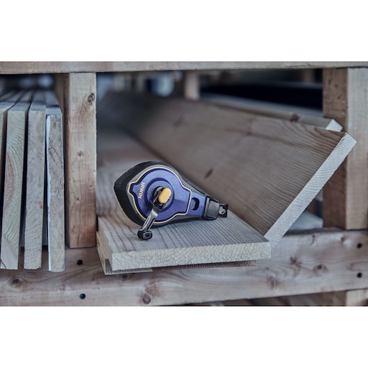 A blue and yellow IRWIN chalk line reel is placed on a wooden plank shelf among stacked lumber boards.